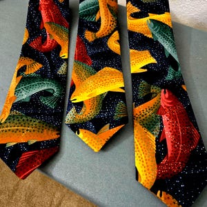 May include: Three neckties with a repeating pattern of colorful fish in shades of red, yellow, and green against a dark blue background with white dots. The ties are arranged on a light blue surface.