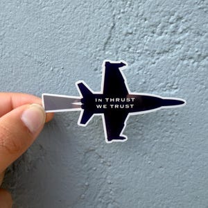 May include: A sticker featuring a stylized black jet with a white and gray exhaust trail. The text "IN THRUST WE TRUST" is centered on the jet's body. The sticker is held against a textured gray wall.