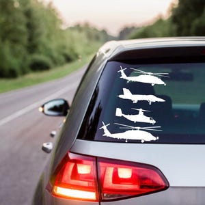 Pode incluir: Um autocolante de vinil branco aplicado no vidro traseiro de um carro, apresentando quatro silhuetas distintas de aeronaves militares. O design inclui dois helicópteros, um jato de combate e uma aeronave de rotores basculantes.