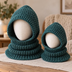May include: Two dark teal crochet balaclavas, one larger and one smaller, each featuring a textured stitch pattern, a fitted hood, and a ribbed neck section. These head coverings are displayed on two plain, off-white mannequin heads.