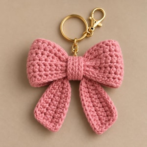 May include: A dusty pink crochet bow keyring featuring a textured yarn construction with a central knot detail. The bow is attached to a gold-toned metal split ring and a lobster claw clasp, providing a secure attachment point for keys or accessories.
