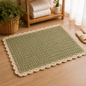 May include: A rectangular crochet bath mat in a sage green color featuring a textured grid pattern throughout the center. The mat is finished with a decorative cream-colored scalloped border that runs along all four edges of the piece.