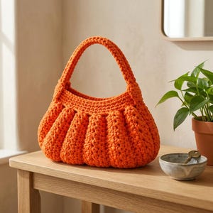 May include: A bright orange crochet handbag featuring a rounded, pleated body and a single integrated handle. The bag is constructed with a thick, textured yarn in a consistent stitch pattern, creating a soft, curved silhouette with vertical ridges.