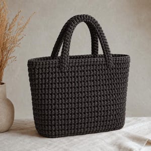 May include: A dark charcoal gray crochet handbag featuring a structured rectangular body and two matching top handles. The bag is crafted with a dense, uniform stitch pattern, creating a textured surface throughout the exterior and handles.