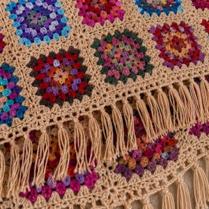 Puede incluir: Una manta de ganchillo con un patrón de cuadrados de la abuela multicolores en tonos morado, azul, rojo, naranja y verde, unidos por un borde de hilo beige claro. El borde está rematado con una fila de borlas de hilo beige claro.