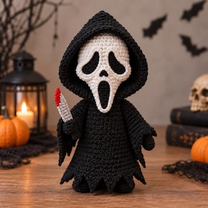 May include: A crocheted figure wearing a black hooded robe and a white mask with black eye and mouth cutouts. The figure holds a small gray knife with a red tip in its right hand. The construction features visible yarn stitches throughout.