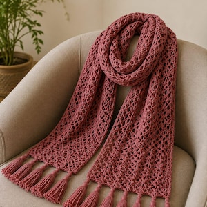 May include: A dusty rose scarf featuring an intricate openwork crochet pattern throughout the length of the fabric. The scarf is finished with multiple matching tassels attached to both ends, creating a textured and uniform appearance.