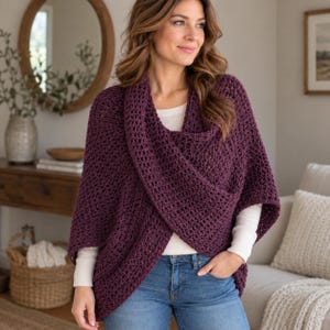May include: A deep plum-coloured, open-front crochet wrap featuring a textured, loose-knit pattern. The garment has wide sleeves and a draped, overlapping front design, worn over a plain white long-sleeve shirt and blue denim jeans.