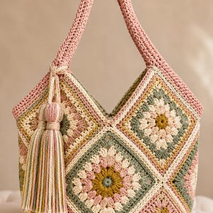 May include: A crochet handbag constructed from multiple square motifs in shades of dusty pink, sage green, and mustard yellow. The bag features a single thick handle and a large, multi-colored yarn tassel attached to the side with a braided cord.
