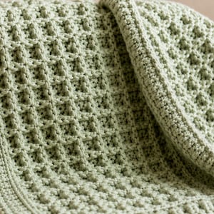 May include: A sage green crocheted blanket featuring a textured waffle stitch pattern with a solid border, accompanied by a small cream-colored crocheted rabbit toy and a woven wicker basket with two handles, all arranged on a flat surface.