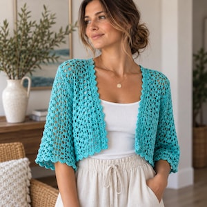 May include: A bright turquoise crochet cardigan featuring an open-knit pattern with scalloped edges along the front opening and sleeves. The garment is worn over a simple white tank top and light-colored drawstring trousers, showcasing a textured design.