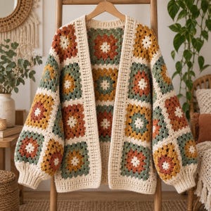 Op de afbeelding: Een vest met lange mouwen en open voorzijde, gemaakt van gehaakte granny squares in tinten crème, mosterdgeel, saliegroen en oranje. Het kledingstuk heeft een herhalend bloemmotief in elk vierkant en een effen, geribbelde crèmekleurige boord.