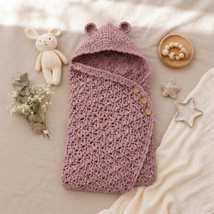 Può includere: Una coperta per neonati all'uncinetto color rosa polvere con cappuccio dotato di due piccole orecchie arrotondate. La coperta presenta un motivo a punto stella testurizzato ed è chiusa lateralmente da tre bottoni in legno chiaro.