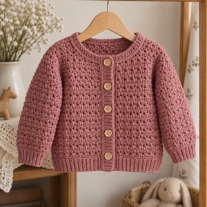 May include: A dusty rose long-sleeve cardigan featuring a textured crochet stitch pattern, a round neckline, and five light-coloured wooden buttons down the centre front. The garment has ribbed cuffs and a ribbed hem, hanging on a wooden hanger.