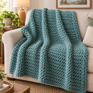 Peut inclure: Un plaid en tricot épais de couleur bleu sarcelle unie avec un motif de fil texturé et volumineux. La couverture est drapée sur le dossier et l'assise d'un canapé de couleur claire, mettant en valeur les boucles denses du tricot.