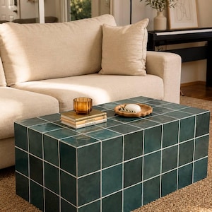 May include: A teal tiled coffee table with white grout lines sits on a woven rug. The rectangular table holds books, a small candle, and a wooden dish with soap. A beige sofa is in the background. The table is approximately 120cm long.