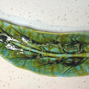 May include: A ceramic leaf-shaped dish with a glossy finish. The dish features a blend of green, blue, and black colors, with a textured surface. The design resembles a stylized leaf, perfect for serving or display.