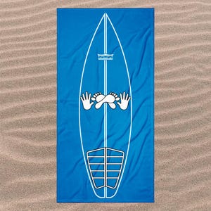 Op de afbeelding: Een rechthoekige strandhanddoek in een levendige blauwe kleur met een grote witte omtrek van een surfplank. De surfplankafbeelding bevat een centrale verticale lijn, een klein kroonicoon, een paar handen, voeten en een traction pad.