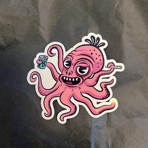 以下が含まれることがあります： 大きな目と広い口を持つピンク色の漫画のタコが描かれたダイカットステッカー。タコは片方の触手で文字Bが書かれた小さな青い缶を持ち、もう片方の触手で細い灰色のタバコを持っており、白い縁取りが施されています。