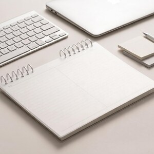 May include: A white spiral-bound notebook with lined pages, featuring a date field at the top left corner. The notebook is positioned next to a silver wireless keyboard, a laptop, a stack of square sticky notes, and a silver pen on a flat surface.