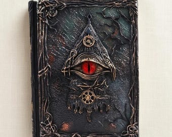 Puede incluir: Un diario rectangular con una cubierta oscura y texturizada que presenta un emblema triangular central con un ojo rojo prominente. El borde está decorado con patrones en relieve de enredaderas en bronce y negro, creando un aspecto.