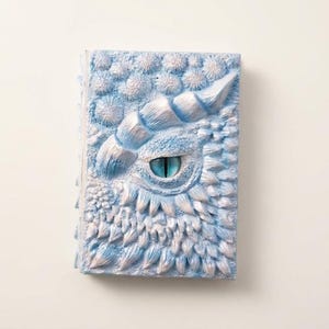 Op de afbeelding: Een rechthoekig notitieboek met een gestructureerde, lichtblauwe en witte kaft met een gebeeldhouwd drakenoog in het midden. De kaft is versierd met verhoogde, schubachtige patronen en een opvallende hoornvorm boven het centrale blauwe oog.