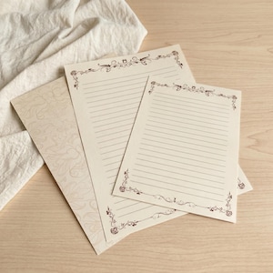 May include: Two sheets of cream-coloured stationery with dark brown horizontal lines and decorative floral scrollwork borders at the top and bottom corners. The paper features a subtle, repeating watermark pattern of stylised leaves and vines.
