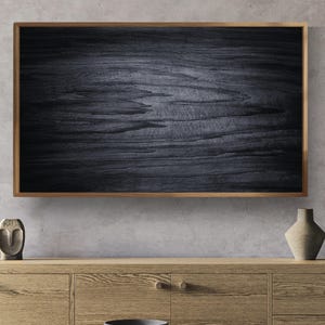 Könnte beinhalten: Ein rechteckiges Wandbild mit einer dunkel anthrazitfarbenen Holzmaserung, eingefasst in einen hellbraunen Holzrahmen. Das Kunstwerk zeigt horizontale, texturierte Linien, die das natürliche Aussehen von verwittertem Holz nachahmen.