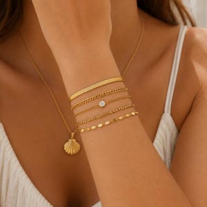 Op de afbeelding: Een set van vijf goudkleurige armbanden om de pols, bestaande uit een platte visgraatketting, een gourmetketting, een fijne ketting met een enkele ronde doorzichtige steen, een getextureerde schakelketting en een ketting met kleine ovale schakels, plus een schelphanger.