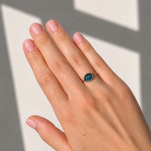 Pode incluir: Um anel de ouro com uma pedra preciosa azul de formato oval e superfície lisa, montada numa banda metálica simples. A pedra tem um aspeto azul profundo e translúcido, fixada numa fina moldura dourada sobre um aro de anel polido e fino.