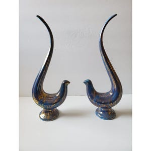 May include: A pair of tall, stylised bird figurines crafted from ceramic with a glossy finish. The birds feature a deep blue base colour with irregular, metallic gold paint splatters and streaks across their curved bodies and circular bases.