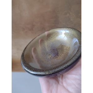 May include: A small, circular glass bowl featuring a translucent, smoky brown tint. The interior surface is decorated with a dense, shimmering gold-coloured flecked pattern that is concentrated around the rim and fades towards the centre.