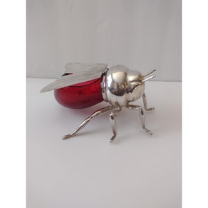 May include: A decorative insect-shaped container featuring a polished silver-toned metal head, legs, and wings. The body of the insect is crafted from translucent deep red glass, providing a contrast between the metallic and glass elements.