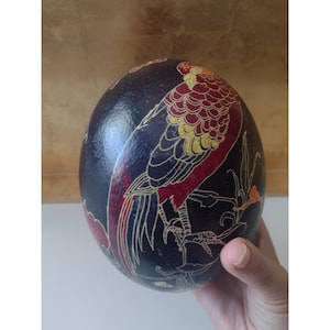 Op de afbeelding: Donkerblauw, eivormig decoratief object met een gedetailleerde illustratie van een vogel. De vogel heeft rode, gele en witte accenten. Het kunstwerk is geplaatst tegen een donkere achtergrond met bloemmotieven.