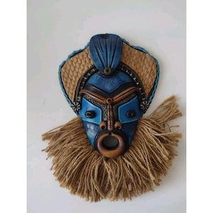 May include: A blue and brown tribal mask with a textured design. The mask features a blue face with brown accents, a woven straw-like background, and a jute beard. The mask has a circular wooden ring and is designed for wall hanging.