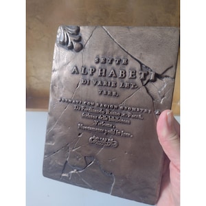 May include: A rectangular bronze-colored plaque with a cracked stone texture featuring the embossed text SETTE ALPHABETI DI VARIE LETTERE, FORMATI CON RAGION GEOMETRY, Da Ferdinando Ruano, scrittor della biblioteca Vaticana, Nuovamente posti in luce.