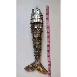 May include: A decorative fish sculpture covered in iridescent abalone shell scales, featuring a layered design and a prominent tail. A pink measuring tape placed alongside the object indicates a total length of approximately 15 inches.