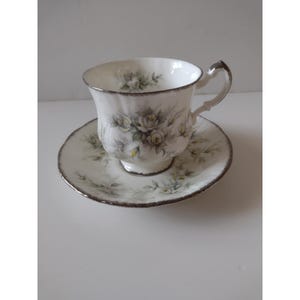 Pode incluir: Conjunto de chá branco com design floral. A chávena e o pires têm uma borda prateada. O design floral apresenta rosas brancas e amarelas com folhas verdes. A chávena tem uma pega curva.