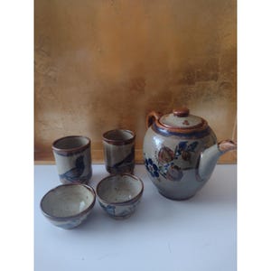 May include: A ceramic tea set consisting of a teapot with a lid and four matching cups. The pieces are light beige with brown speckled details, featuring hand-painted floral motifs and bird illustrations in blue, brown, and orange tones.
