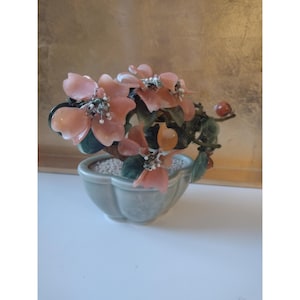 May include: A decorative bonsai tree with peach-colored blossoms and green leaves. The tree is in a light green, scalloped-edge pot. The background is a gold-toned surface.