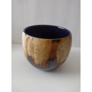May include: A rounded ceramic bowl featuring a deep navy blue interior and a textured exterior with a mottled tan and brown glaze that drips down towards the base, revealing the dark blue colour underneath the earthy, light-coloured surface.