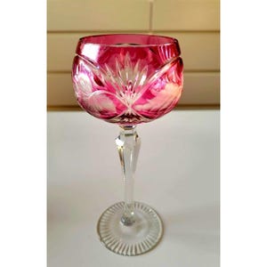 Könnte beinhalten: Ein rubinrotes Kristall-Weinglas mit einem klaren, geätzten Stiel und Fuß. Die Schale hat ein Blumendesign mit weißen Akzenten. Das Glas ist für Wein konzipiert und eignet sich für besondere Anlässe.