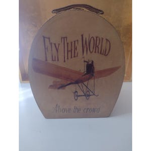 Peut inclure: Une valise ovale de couleur beige avec une poignée sur le dessus. La face avant présente une illustration marron foncé d'un biplan vintage avec le texte "FLY THE WORLD" en grandes lettres au-dessus et "Above the crowd" en dessous.
