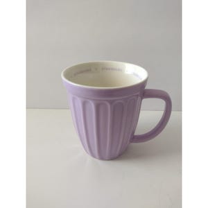 Puede incluir: Una taza de cerámica de color morado claro con una textura acanalada vertical en el exterior. El interior es blanco y presenta la palabra STARBUCKS impresa repetidamente en una fuente morada clara a lo largo del borde interior. Incluye un asa a juego.