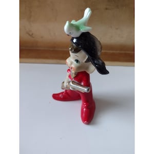 Puede incluir: Figura vintage de un elfo sentado con un atuendo rojo, sombrero negro y un pájaro verde y blanco en la parte superior. El elfo sostiene un instrumento plateado. La figura está sobre una superficie blanca.