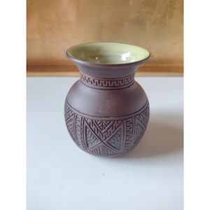 May include: A dark brown ceramic vase with a wide, flared opening and a light green interior glaze. The exterior features a band of geometric patterns around the neck and a series of repeating triangular motifs with textured details.