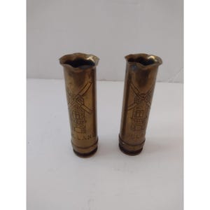 May include: Two cylindrical brass casings with scalloped top edges. Each piece features an etched design of a windmill and the text HOLLAND engraved near the base. The metal has a tarnished, aged appearance with visible vertical lines.