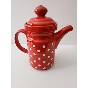 May include: A red ceramic teapot with a lid and a handle. The teapot features white polka dots. The spout is curved, and the handle is a large loop. The teapot is on a white surface.