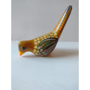 May include: A hand-painted wooden bird figurine featuring a mustard yellow base color with intricate patterns in green, orange, white, and black. The bird has a long, pointed tail, a small orange beak, and detailed feather motifs across its body.