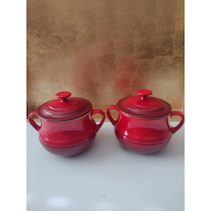 May include: Two matching round red ceramic pots with lids and side handles. Each pot features a glossy finish with a darker red gradient towards the edges and a small knob on the centre of the lid. The brand name Le Creuset is embossed on the lids.